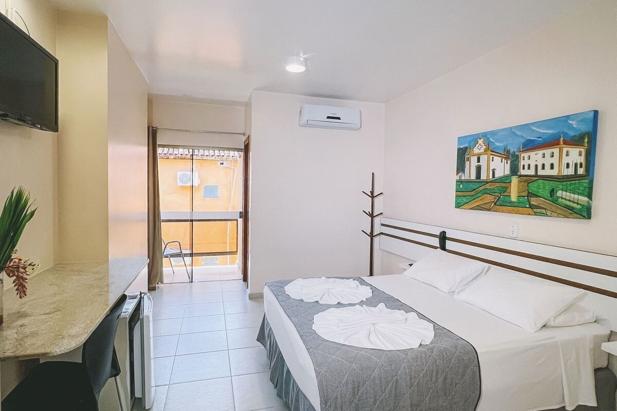 A hotel room with a bed, a desk, a chair, an air conditioning unit, and a painting on the wall.