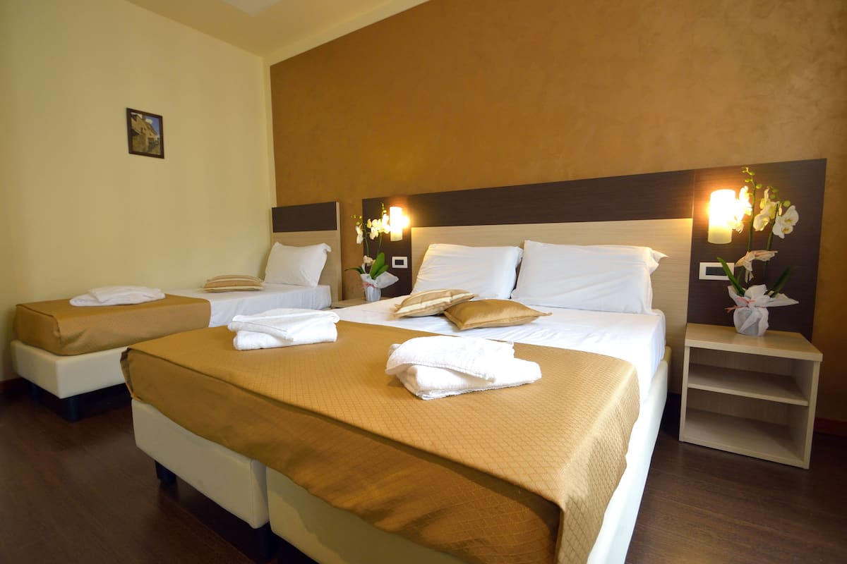 Habitación de hotel con dos camas, cada una con sábanas blancas y una manta marrón. Hay dos mesitas de noche con lámparas y un florero.