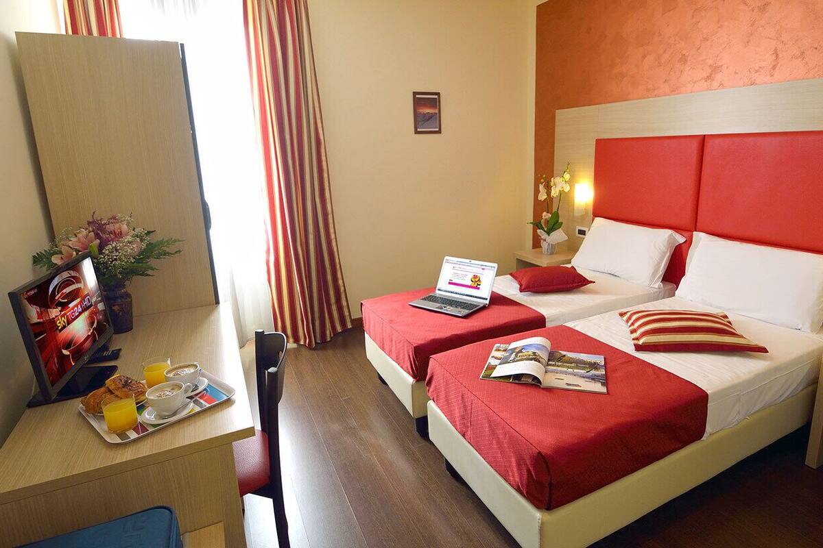 Habitación de hotel con dos camas, un escritorio con una computadora portátil, una mesita con desayuno y un televisor.