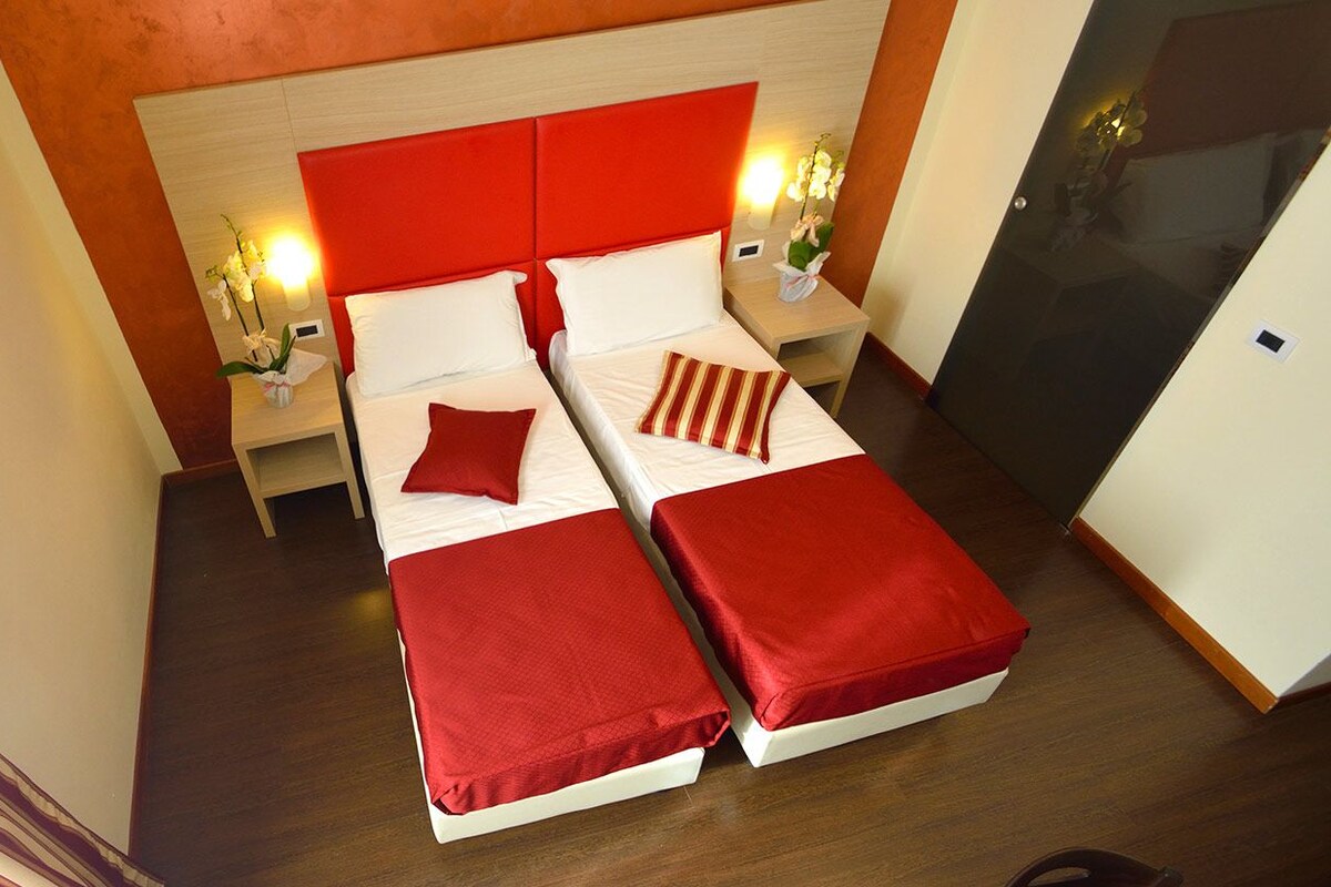 A hotel room with two beds, red bedding, wooden headboard, bedside lamps, and a small table with a vase of flowers.