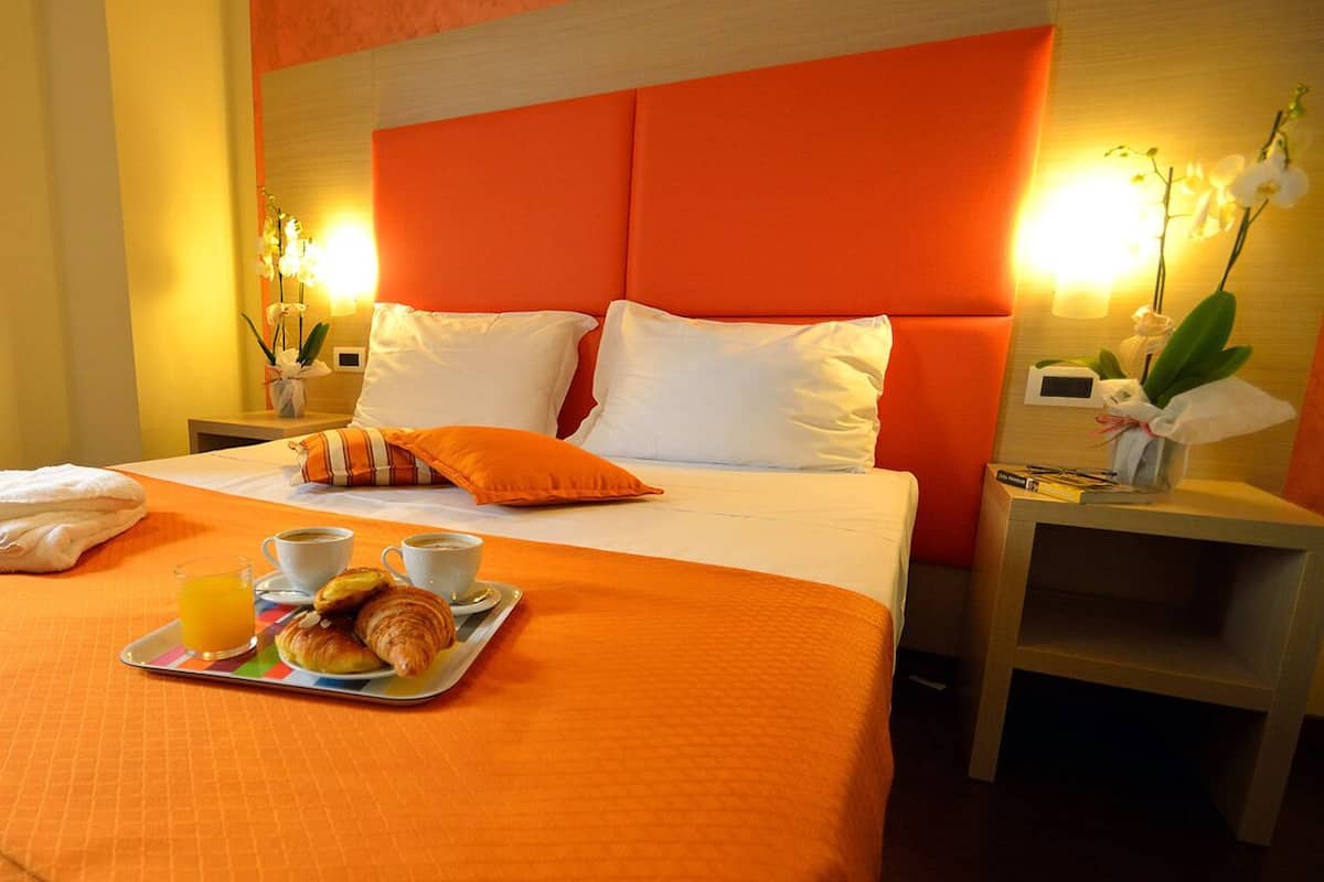 Una habitación de hotel con cama, dos mesitas de noche, una bandeja con croissants y jugo, y un jarrón con flores.