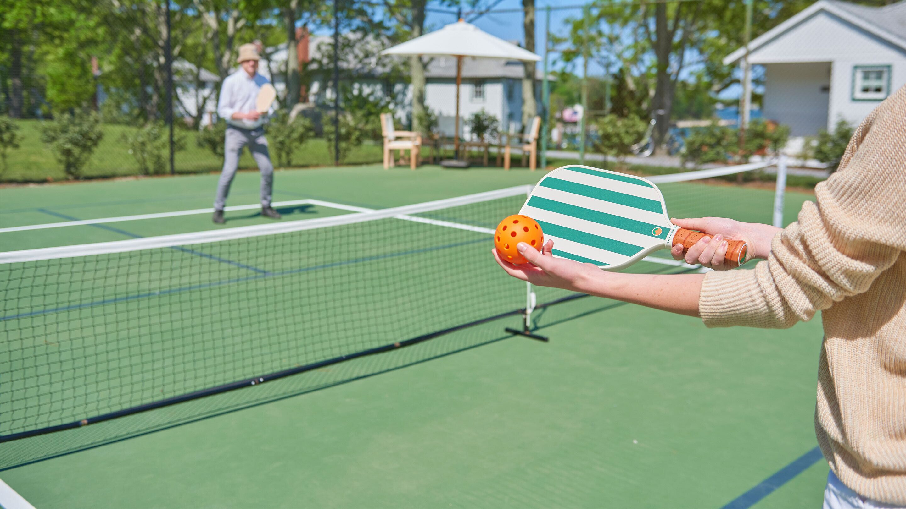 Ein Tennisplatz mit Netz, ein Spieler im Hintergrund und eine Person, die Schläger und Ball hält.