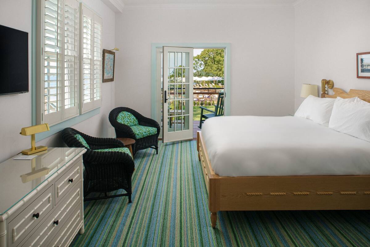 A hotel room with a bed, a dresser, a chair, and a view of the outdoor area through a door.