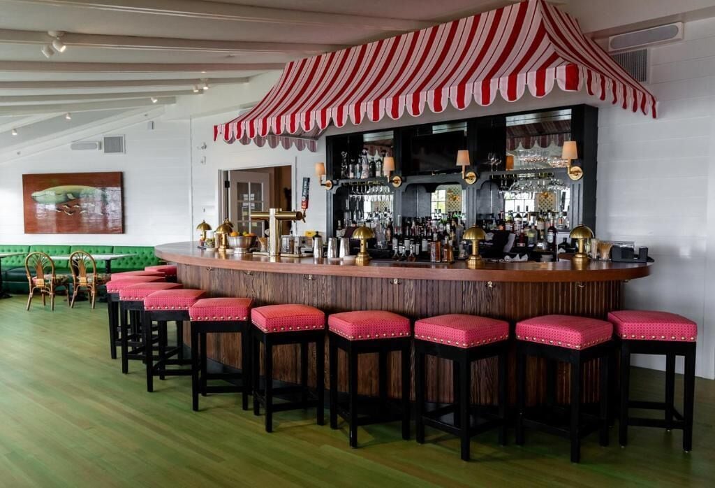 Una zona de bar con un toldo a rayas, una barra con taburetes y un área de estar verde al fondo.
