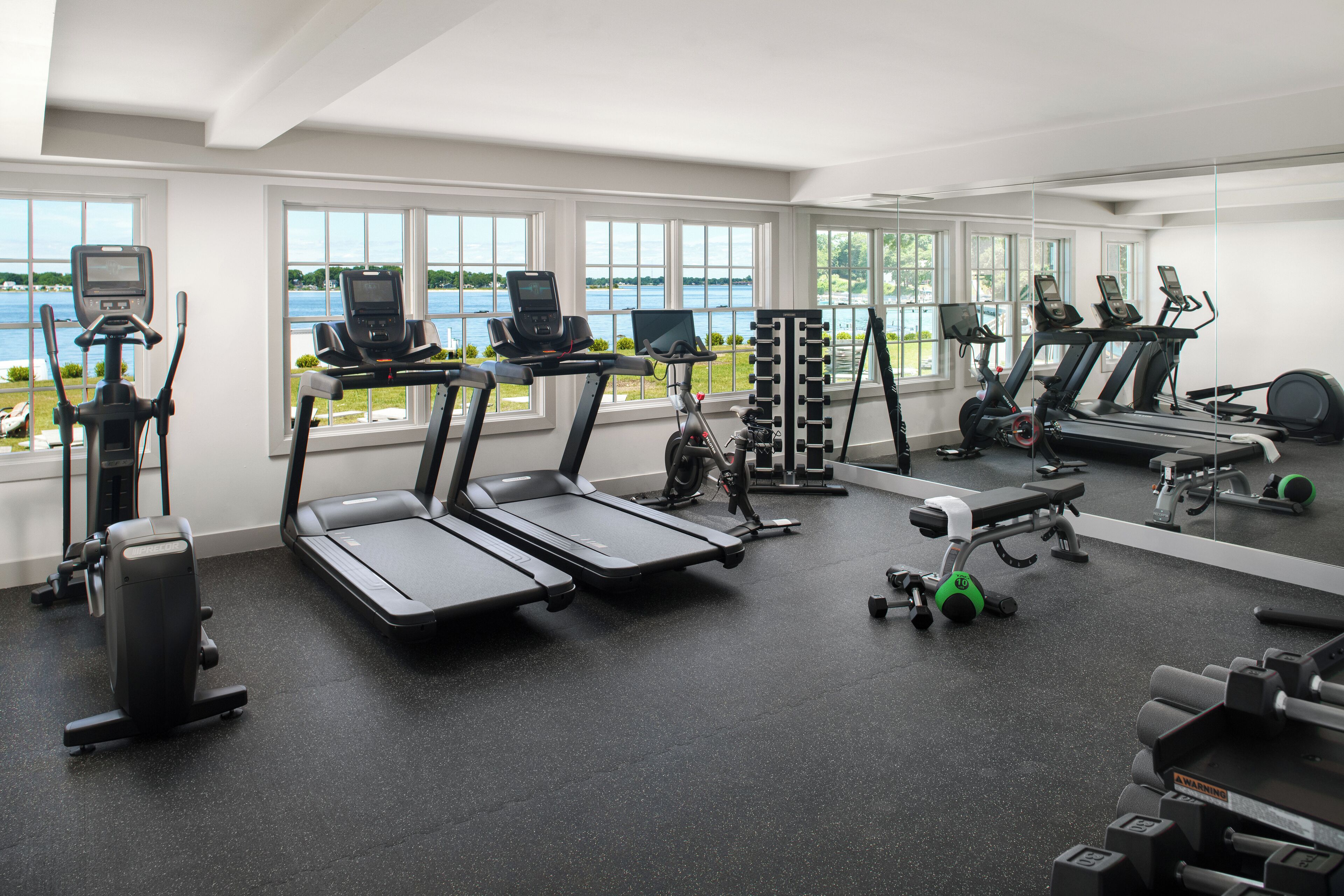 Een sportschool met loopbanden, trainingsfietsen en gewichten, met grote ramen en uitzicht op een meer.