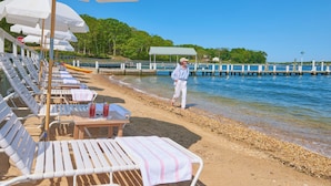 En strand med rader av vita solstolar och parasoller, ett träbord med en drink på och en person som promenerar längs stranden.