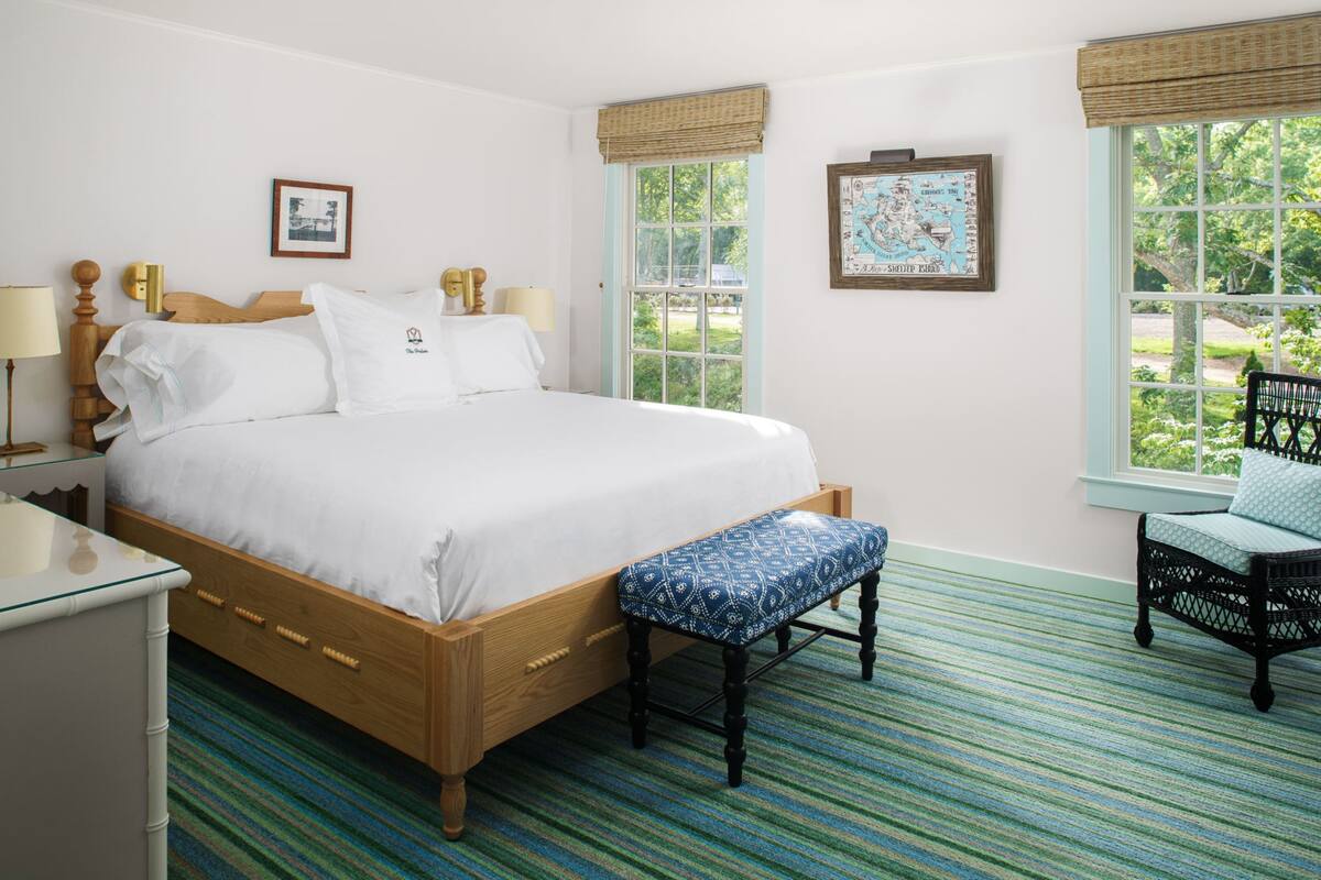 A bedroom with a wooden bed, a bench, a chair, and two windows with views of greenery.