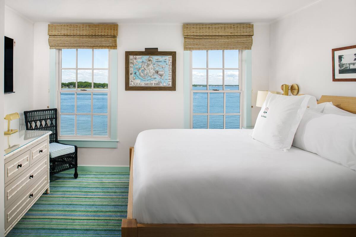 A bedroom with a large bed, two windows with ocean views, a map on the wall, a chair, and a dresser.