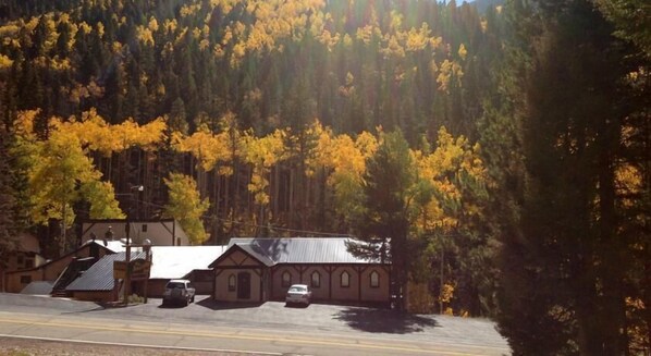 A building with a sloped roof and a small attached structure, surrounded by a forest with autumn foliage.