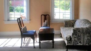 A room with two windows, a chair, a table, and a patterned sofa. - The Teacherage (Sunbury)