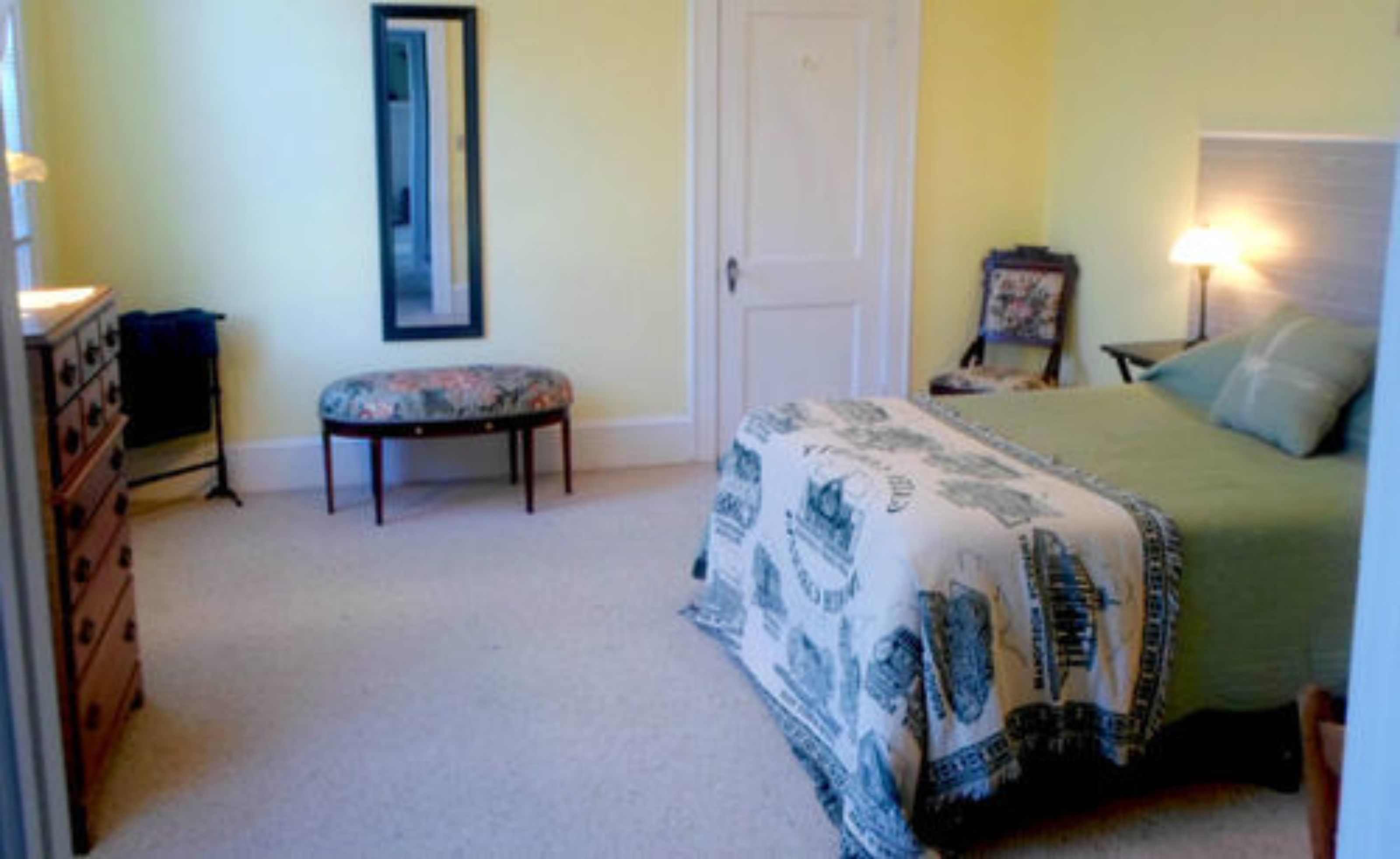 A bedroom with a bed, a dresser, a bench, a door, and a lamp.