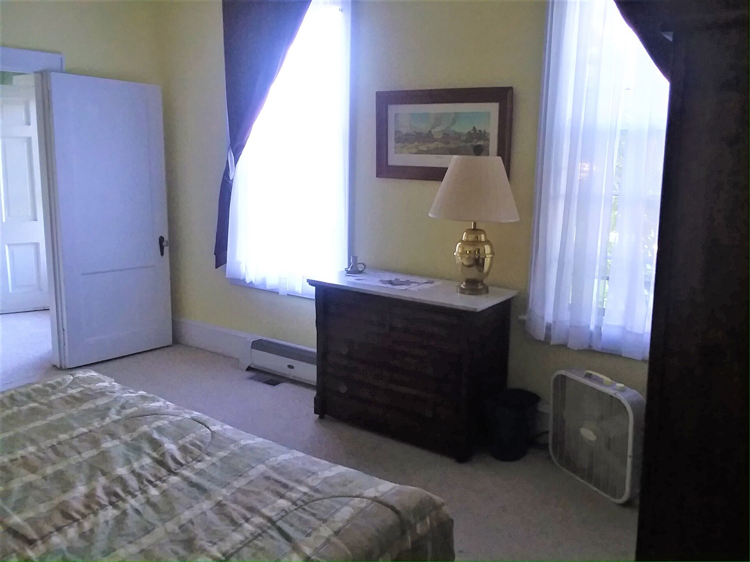 Quarto com cama, cómoda, candeeiro, janela com cortinas e radiador.