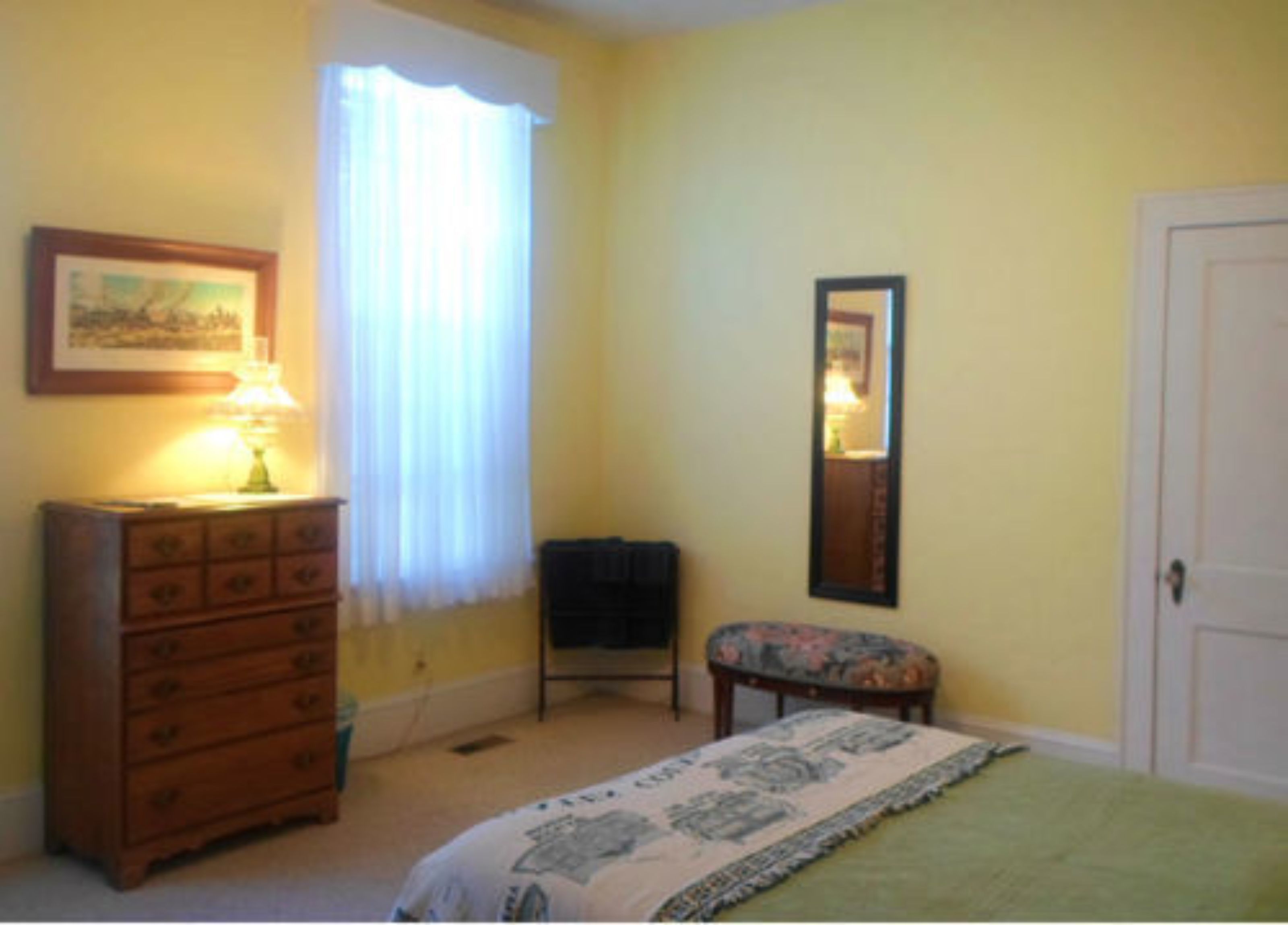 Un dormitorio con un aparador de madera, una silla, una cama con colcha estampada, una ventana con cortinas blancas y un espejo en la pared.