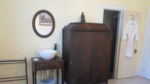 A bedroom with a wooden wardrobe, a bedside table with a bowl, a mirror, and a door leading to another room. - The Teacherage (Sunbury)