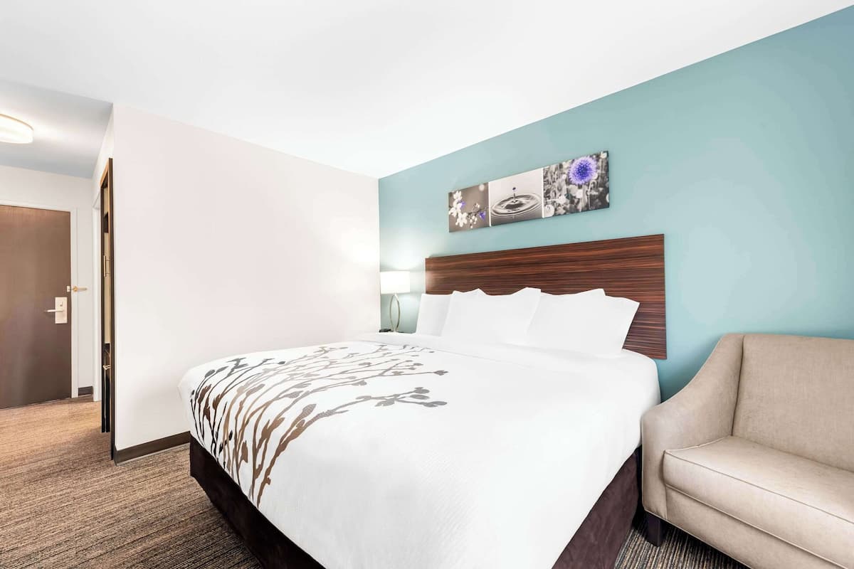 Una habitación de hotel con una cama grande, un cabecero de madera, un sillón color beige y una obra de arte colgada en la pared.