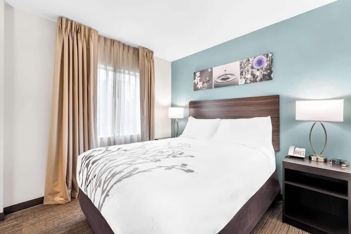 Habitación de hotel con una cama grande, una mesita de noche con lámpara, un reloj de pared y una ventana con cortinas.