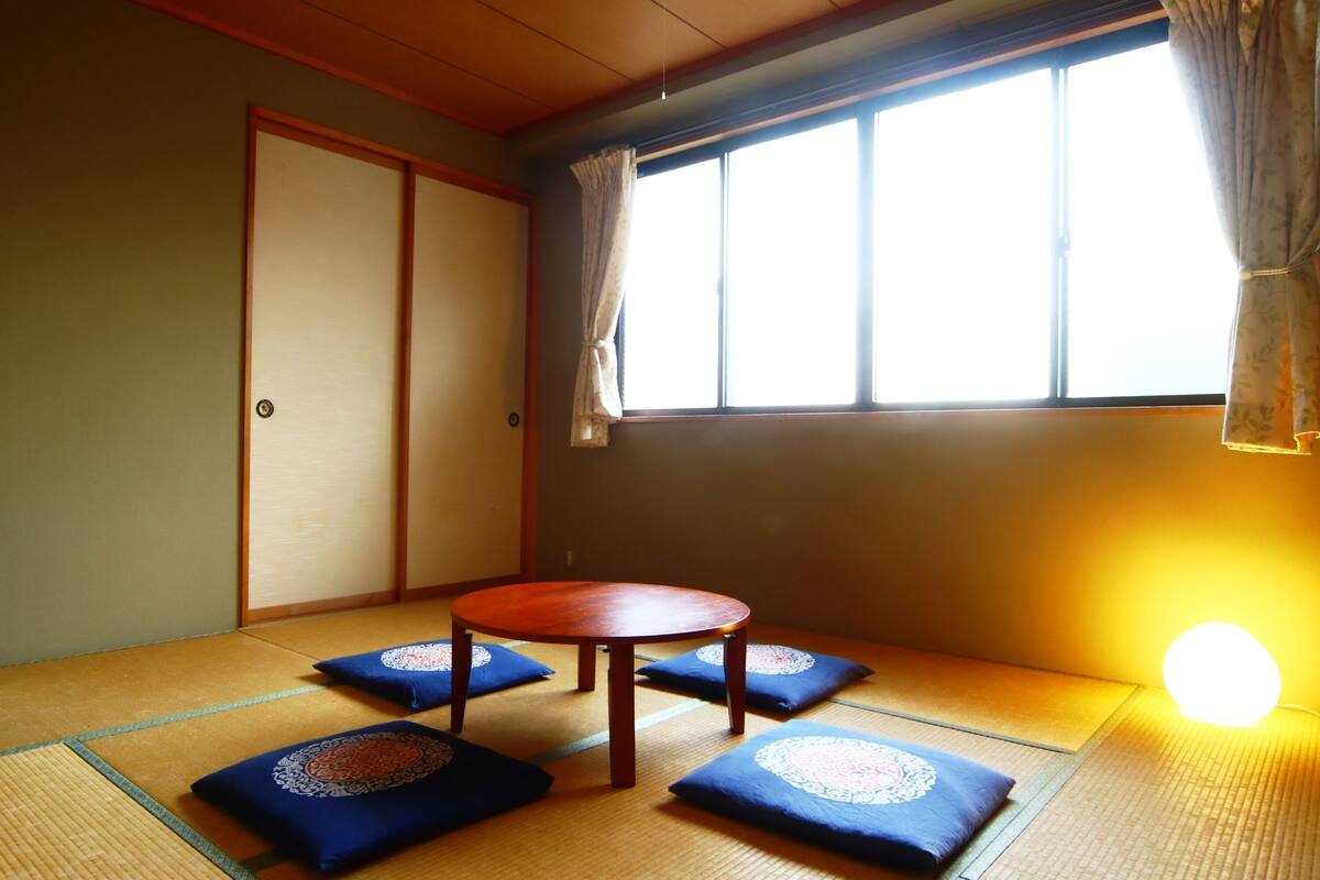 A tatami mat room with a low table and cushions, a sliding door, and a window with curtains.