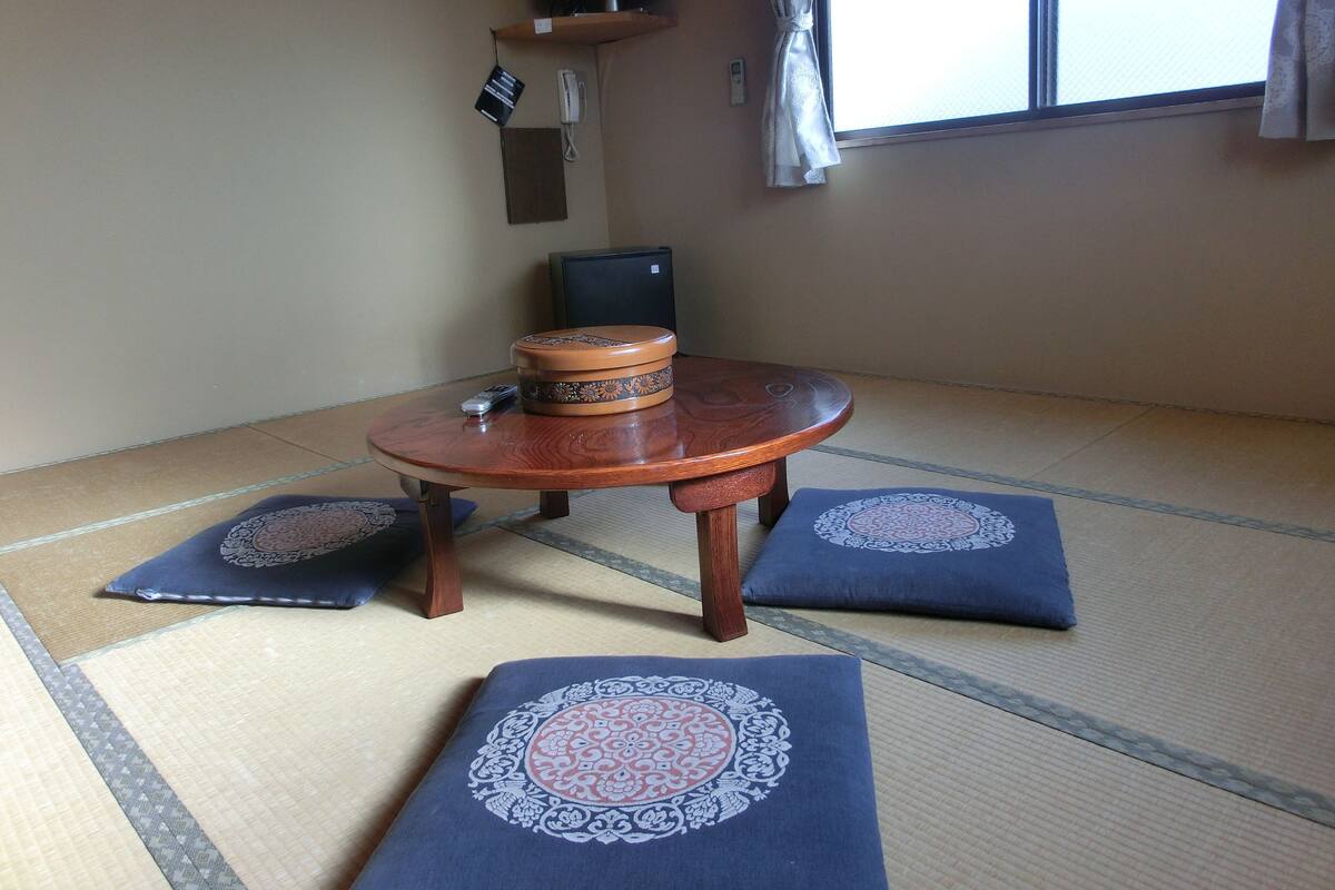 A tatami mat room with a round wooden table, cushions, a speaker, and a television on the wall.