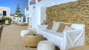 Un área de estar exterior con mobiliario blanco, un muro de piedra de fondo, otomanas de mimbre y un sofá blanco con cojines a rayas.