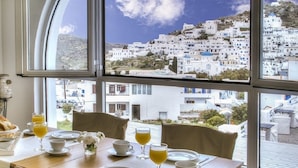Una mesa de comedor dispuesta con alimentos para el desayuno y vasos de zumo de naranja, con vistas a un pueblo blanco con cúpulas azules.