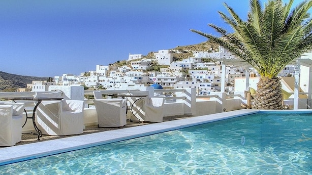 Zona de piscina con sillones blancos y una palmera, con vistas a una colina de edificios blancos.