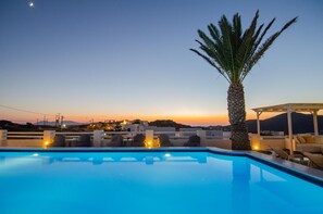 A swimming pool with a palm tree and lounge chairs, overlooking a coastal town at sunset. - Avanti Hotel (Ios)