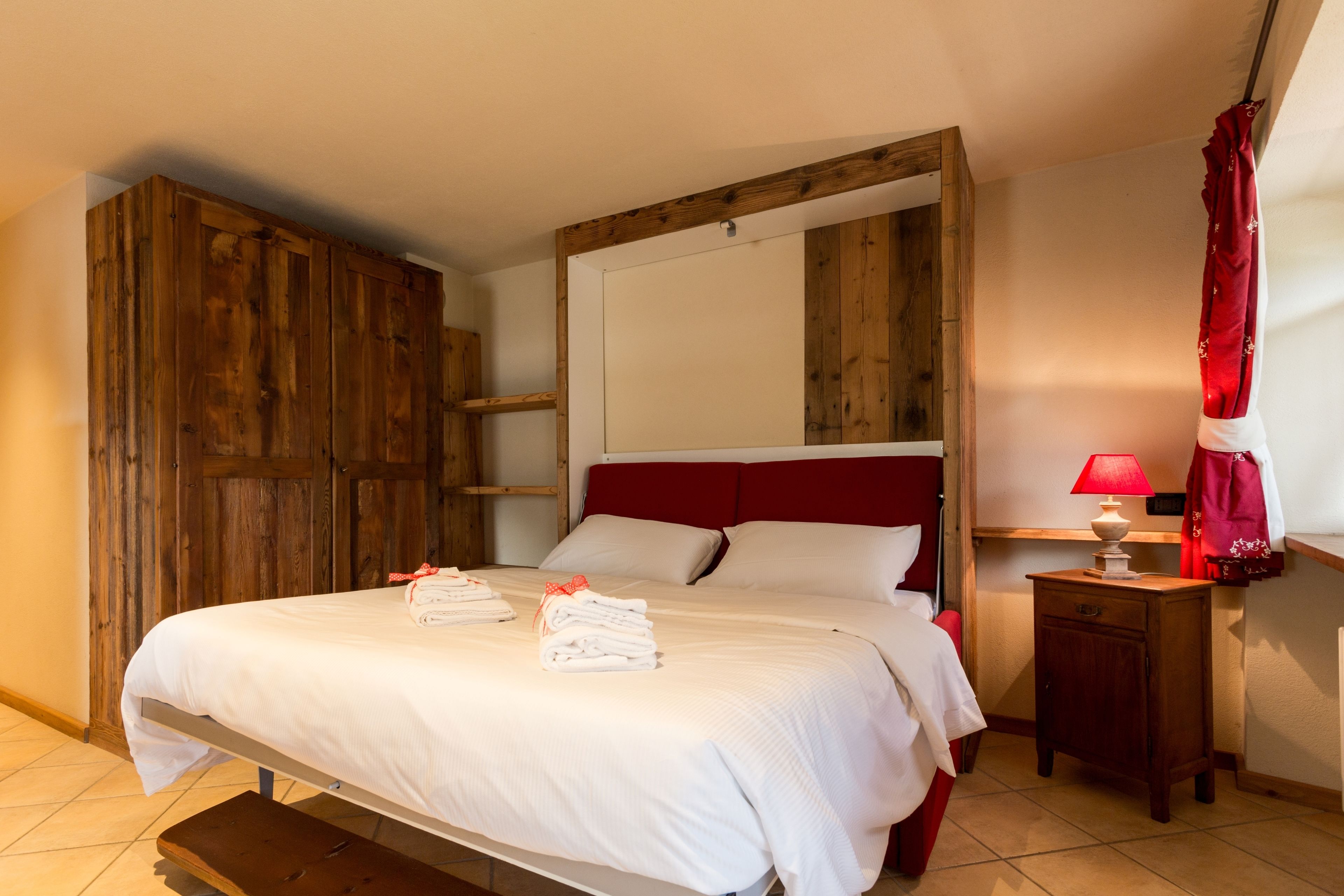 A neatly made bed with white linens and red accents, a wooden wardrobe, a small bedside table with a lamp, and a red curtain.