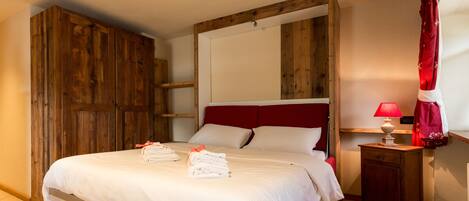 A neatly made bed with white linens and red accents, a wooden wardrobe, a small bedside table with a lamp, and a red curtain.