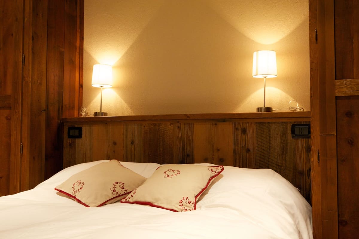 A wooden-framed room with a bed, two bedside lamps, and a wall-mounted thermostat.