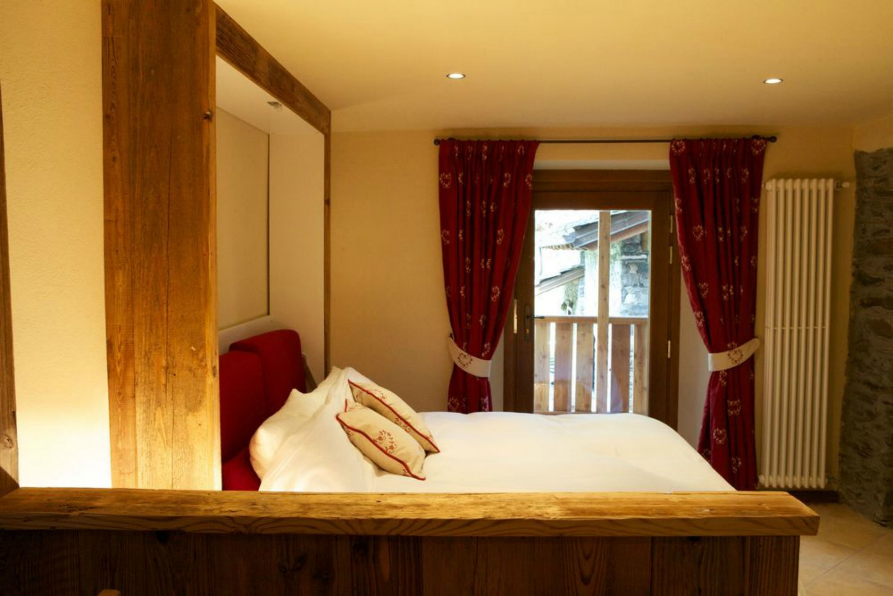 Une chambre à coucher avec une tête de lit en bois, un lit avec une literie blanche et des oreillers rouges, des rideaux rouges et une vue sur un balcon.