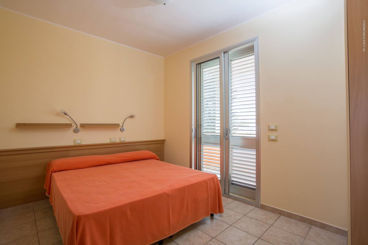 Une chambre à coucher avec un lit, une fenêtre avec des stores, une porte en bois et une étagère fixée au mur.