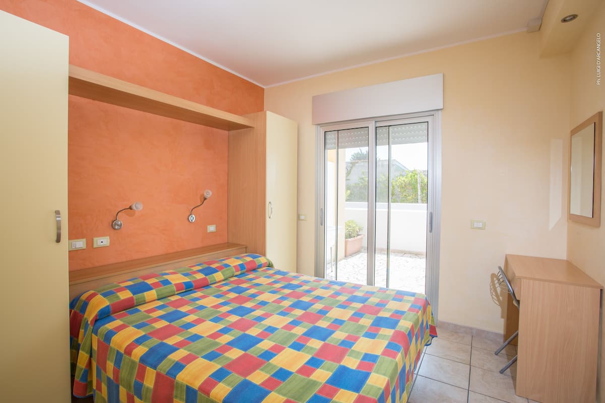 Une chambre avec une couvre-lit à carreaux, une tête de lit en bois, un bureau et une porte-fenêtre coulissante donnant sur un espace extérieur.