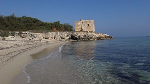 A beach with clear water, a rocky shoreline, and a historic stone building. - Residence Solaris (Melendugno)