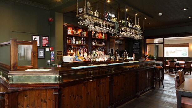 A bar area with a wooden counter, shelves of bottles, and hanging glasses. - The Avenue (Durham)