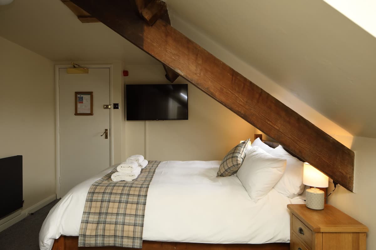 Um quarto com uma cama, televisão fixada na parede, mesa de cabeceira de madeira e uma porta com um mural de avisos.