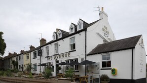 A white building with "The Avenue" sign, outdoor seating, and a green circular sign on the side. - The Avenue (Durham)