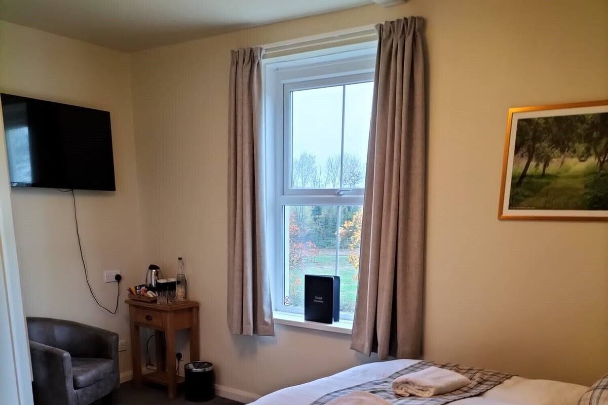Um quarto com cama, mesa de cabeceira com um livro, controle remoto e uma janela com cortinas.