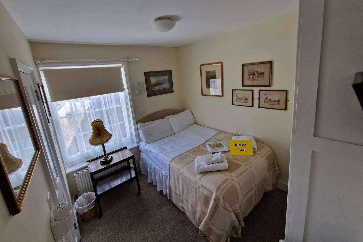 A bedroom with a bed, a bedside table, a lamp, a radiator, and a window with blinds. There are framed pictures on the wall and a sign indicating "ROOM TWO".