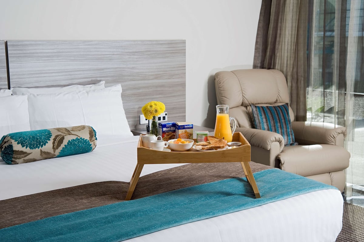 A hotel room with a neatly made bed, a breakfast tray with food and drinks, a comfortable armchair, and a view through the window.