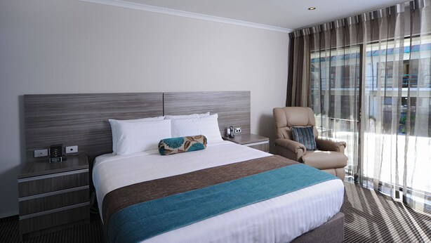 A hotel room with a large bed, a nightstand with a phone and a small table, a chair, and a large window with curtains.