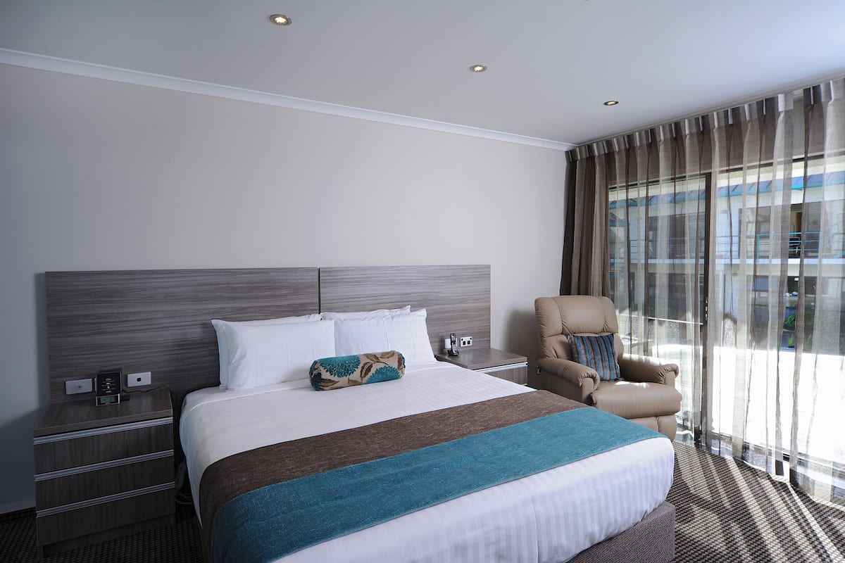 A hotel room with a large bed, a nightstand with a phone and a small table, a chair, and a large window with curtains.