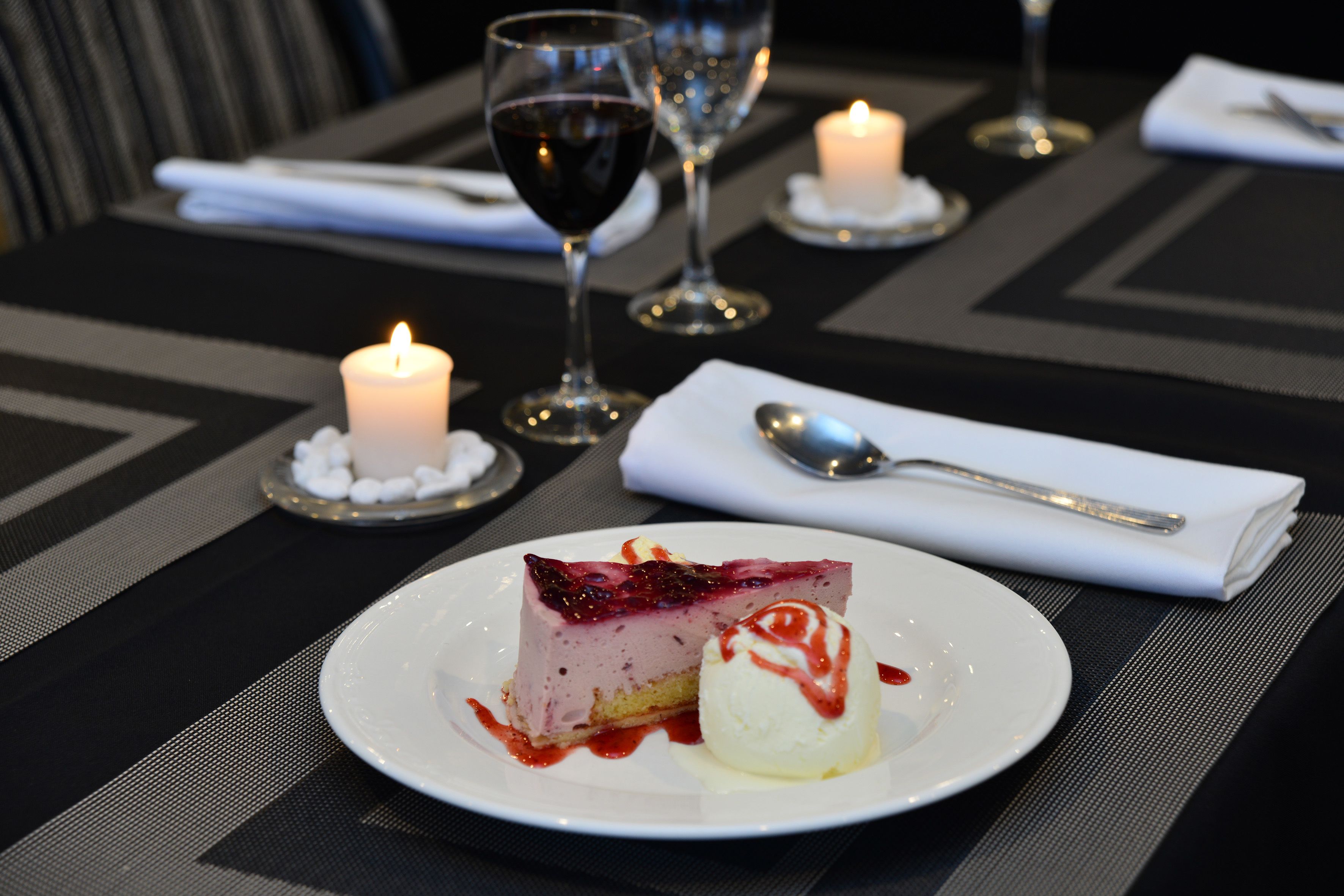 A dessert plate with a slice of cheesecake and a scoop of ice cream, accompanied by a spoon and a napkin on a table with candles and wine glasses.