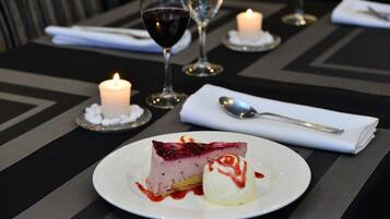 Un plato de postre con una porción de pastel de queso y una bola de helado, acompañado de una cuchara y una servilleta sobre una mesa con velas y copas de vino.