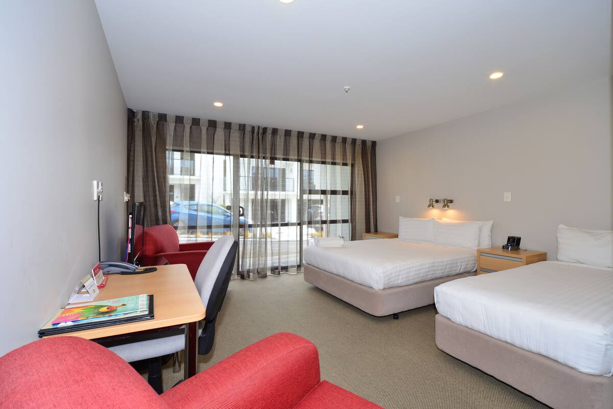 A hotel room with two beds, a desk with a chair, a small table, and a window with curtains.