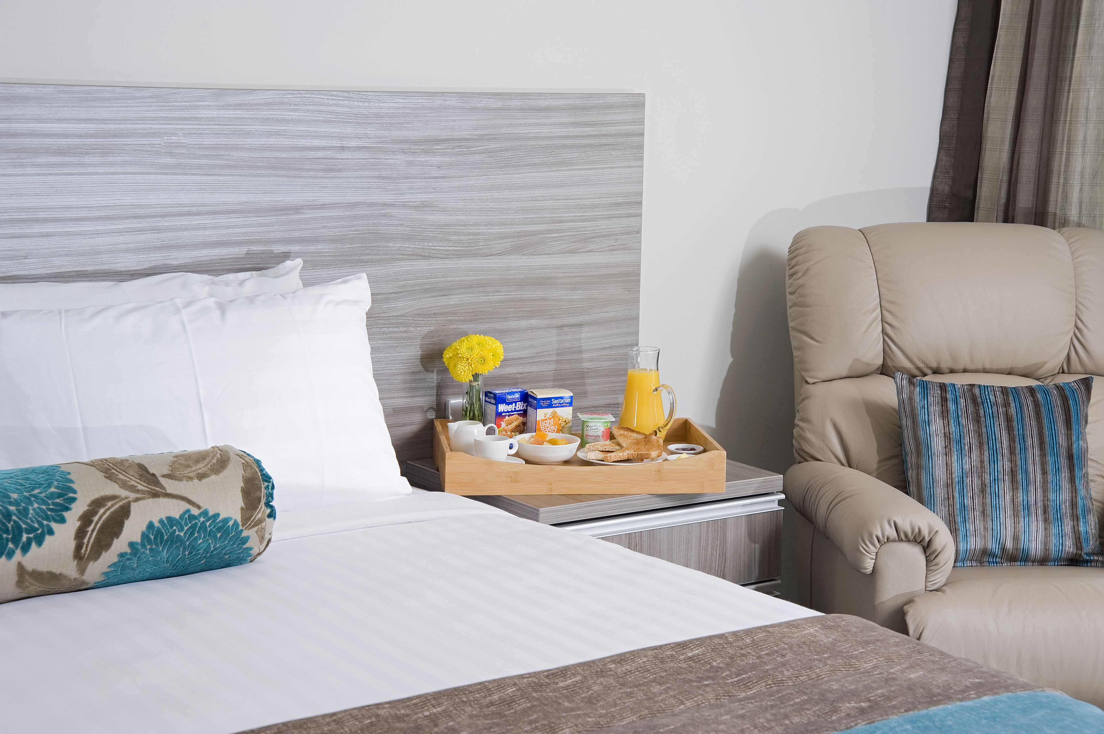 Ein Hotelzimmer mit einem Bett, einem Nachttisch mit Frühstücksgebäck und -speisen, einem Sessel und einem Kopfteil.