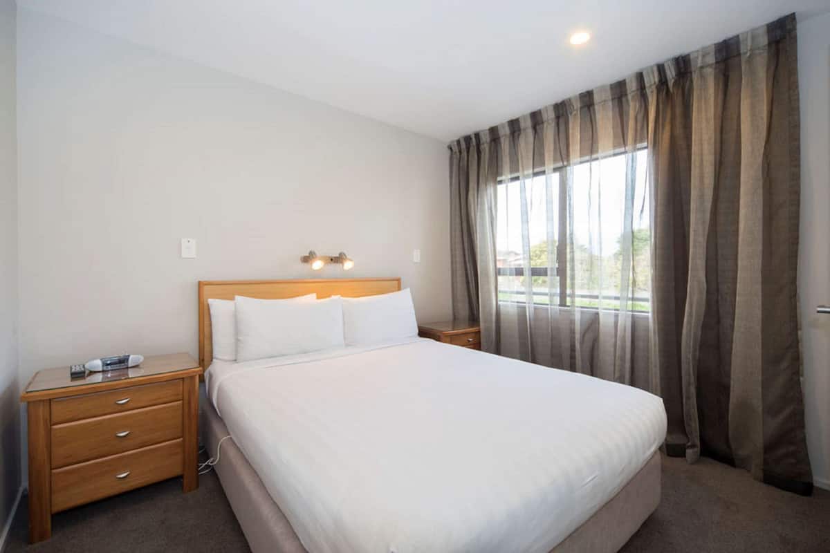A hotel room with a large bed, wooden nightstands, a bedside table, and a window with curtains.