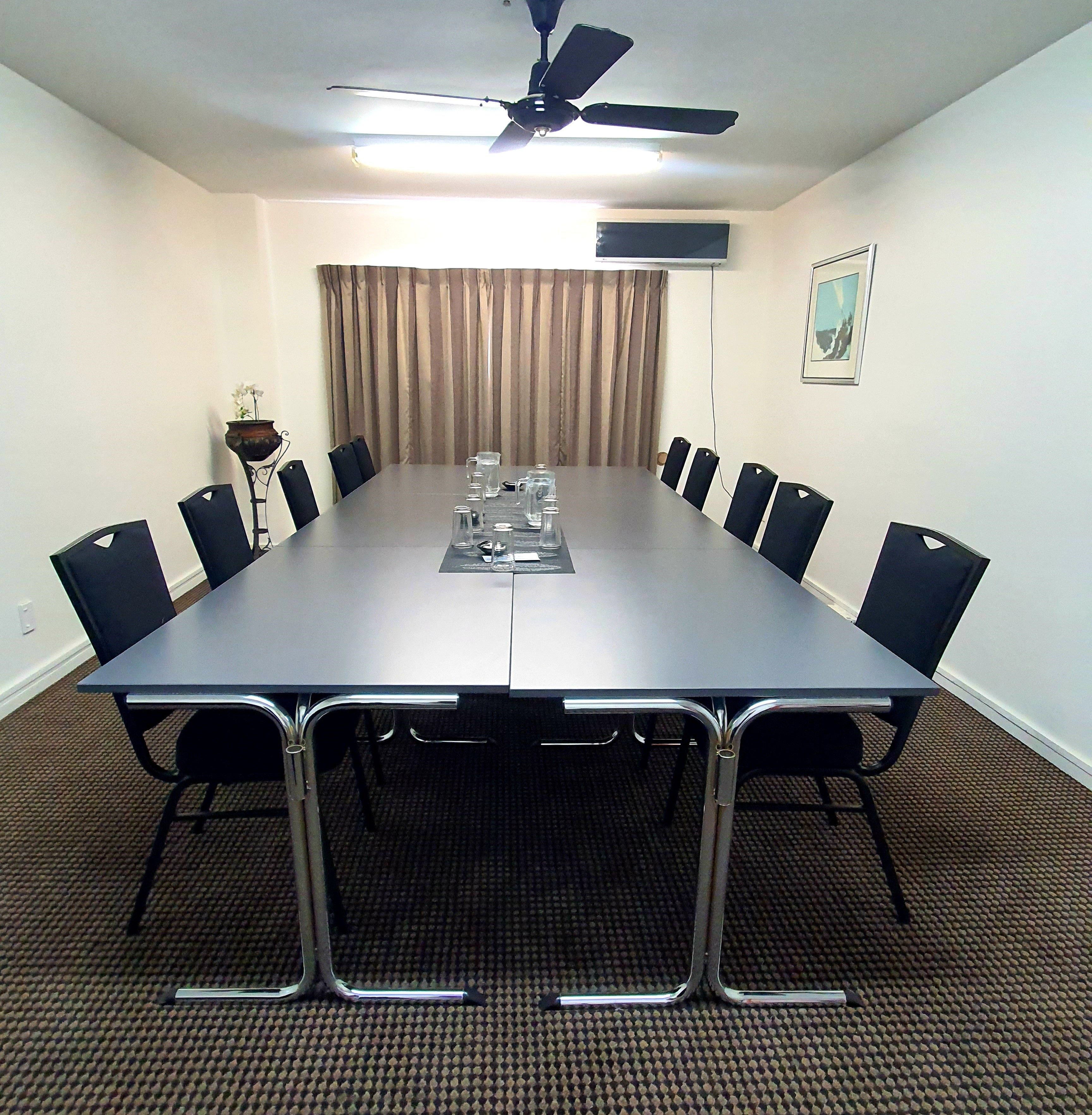Un salón de conferencias con una larga mesa, sillas, un ventilador de techo y una ventana con cortinas.
