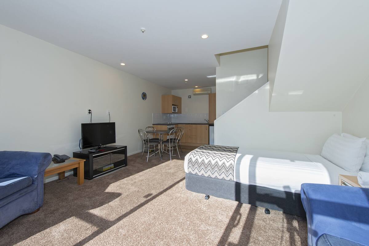 A compact living space with a bed, a sofa, a TV, a small dining table with chairs, and a kitchenette in the background.