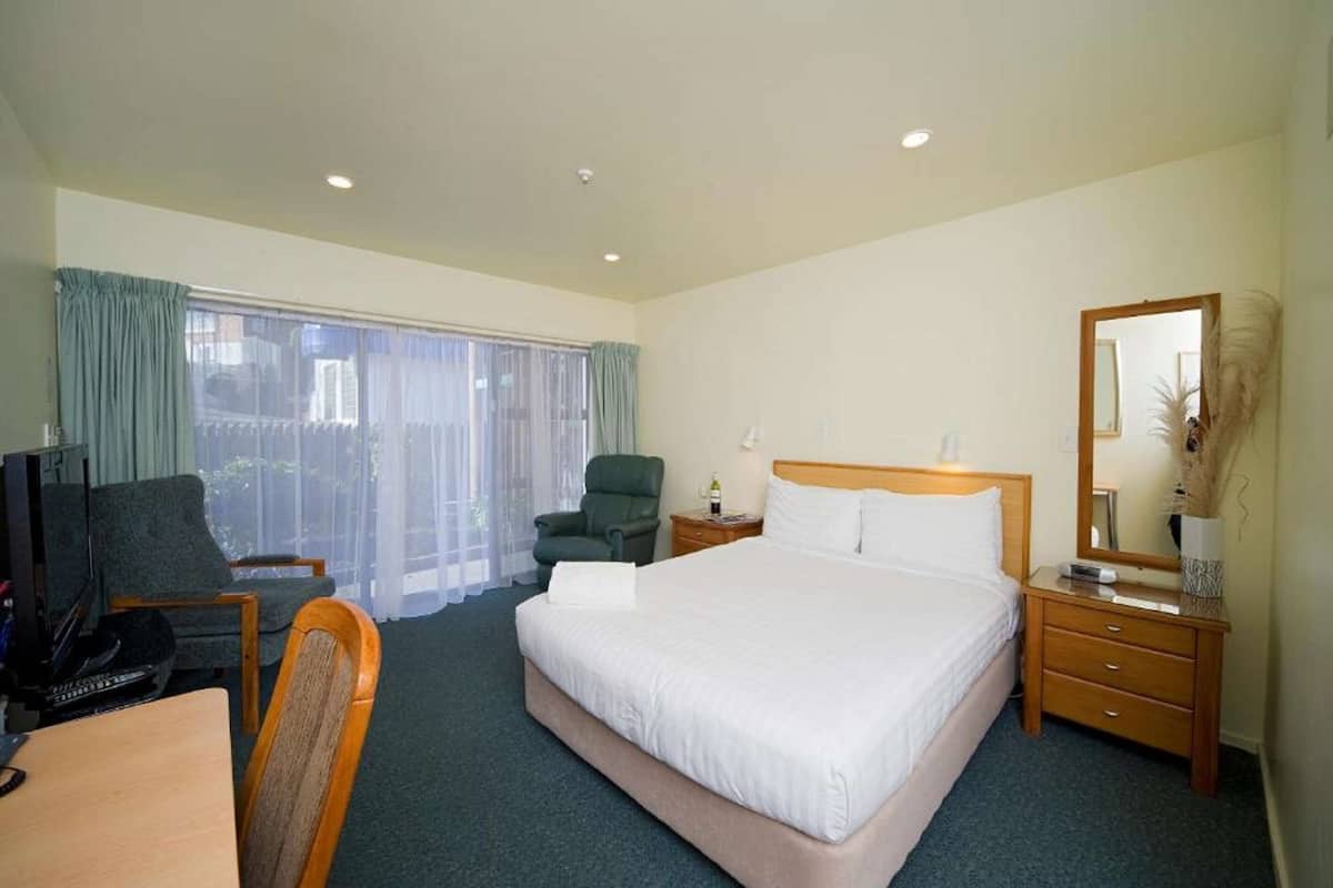 A hotel room with a large bed, a desk with a computer, a chair, a nightstand with a lamp, and a window with curtains.