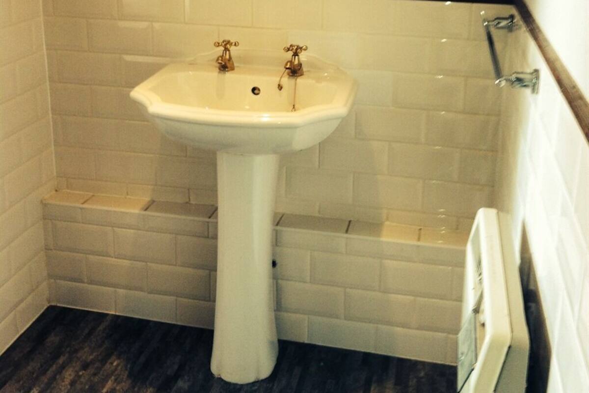 A bathroom with a white pedestal sink, white tiled walls, and a wooden floor.